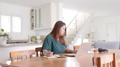 Mujer-Birracial-De-Talla-Grande-Tomando-Notas-Y-Usando-Una-Computadora-Portátil,-Trabajando-Desde-Casa,-Copiando-Espacio,-Cámara-Lenta