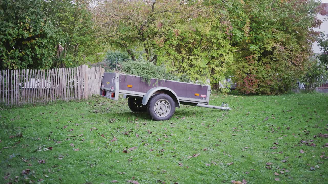 Wide side view of a trailer in a traditional German garden, fully visible and surrounded by greenery, filled with tree and shrub cuttings, capturing the full context of seasonal garden cleanup