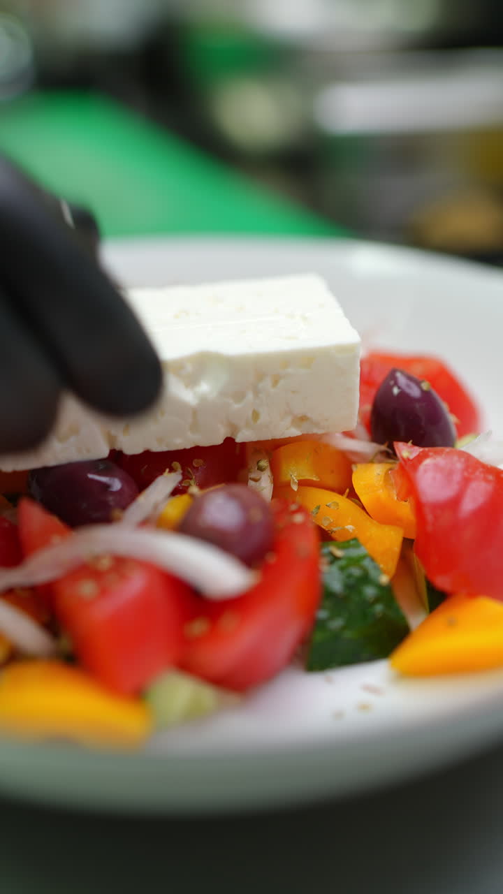 Vertical footage Gloved hands arranging feta block atop vibrant Greek salad featuring ripe tomatoes, crisp cucumbers, red onions, kalamata olives, green peppers