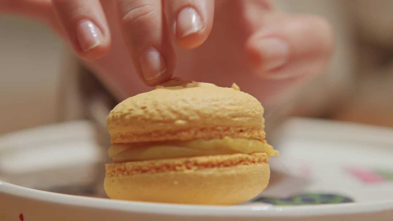 Close up of hand gently pressing down on top of yellow macaron focusing on texture and detail. scene is set indoors with soft lighting showing interaction between human fingers and baked
