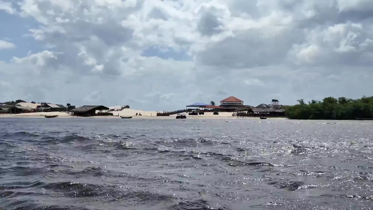 Toàn cảnh từ một chiếc thuyền đang di chuyển trên mặt nước gợn sóng về phía bờ sông cát với những tòa nhà mộc mạc, thuyền và cồn cát ở Maranhao, Brazil.