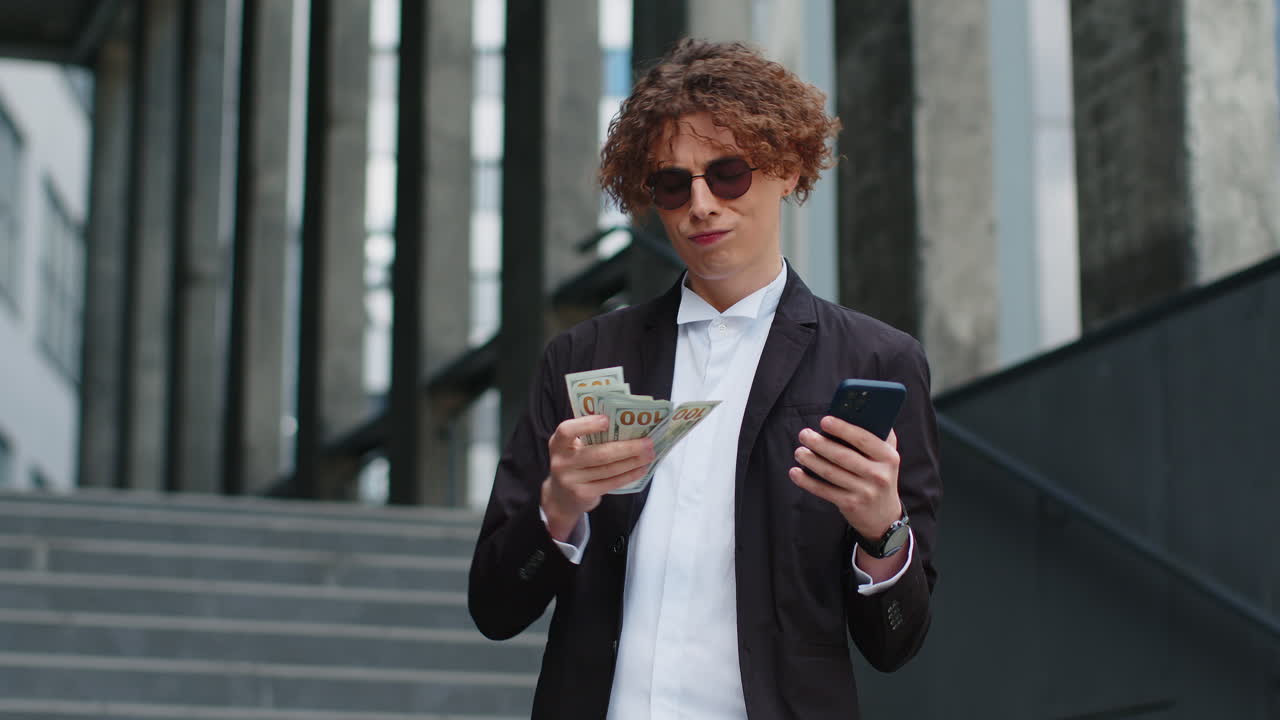 Businessman standing on city street showing winning earnings cash money with mobile phone in hands