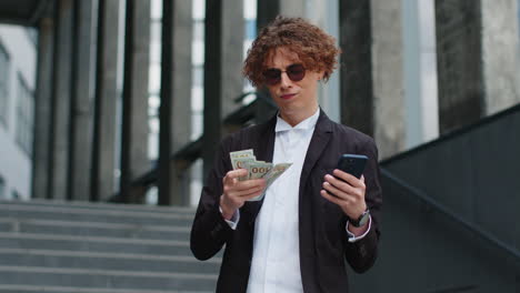 businessman standing on city street showing winning earnings cash money with mobile phone in hands