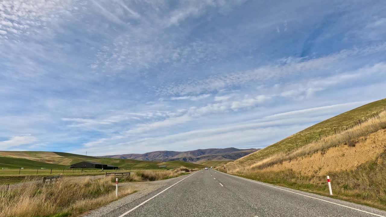 A serene drive along a winding road in Queenstown, New Zealand, surrounded by lush hills under a clear blue sky