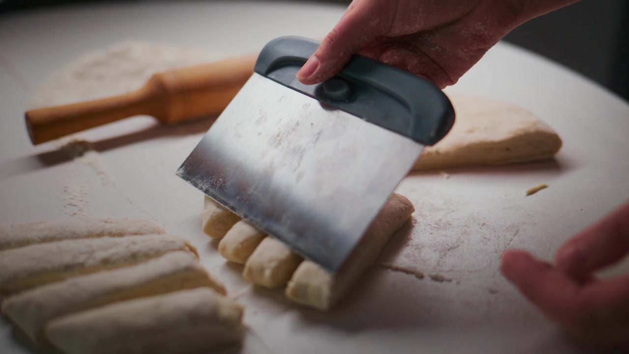 Hände schneiden Teig mit einem Messer. Teigstücke liegen ordentlich angeordnet. Ein Nudelholz liegt in der Nähe auf einer weißen Oberfläche. Die Handlung zeigt die Zubereitung von Brot oder Gebäck. Ideal für Rezepte, Koch-Tutorials