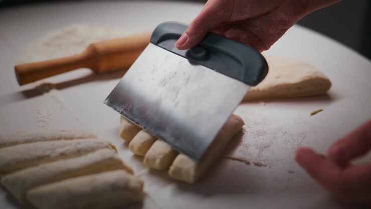 Mani affettano l'impasto con un coltello. I pezzi di impasto sono disposti ordinatamente. Un mattarello riposa nelle vicinanze su una superficie bianca. L'azione mostra la preparazione di pane o prodotti da forno. Ideale per ricette, tutorial di cucina.
