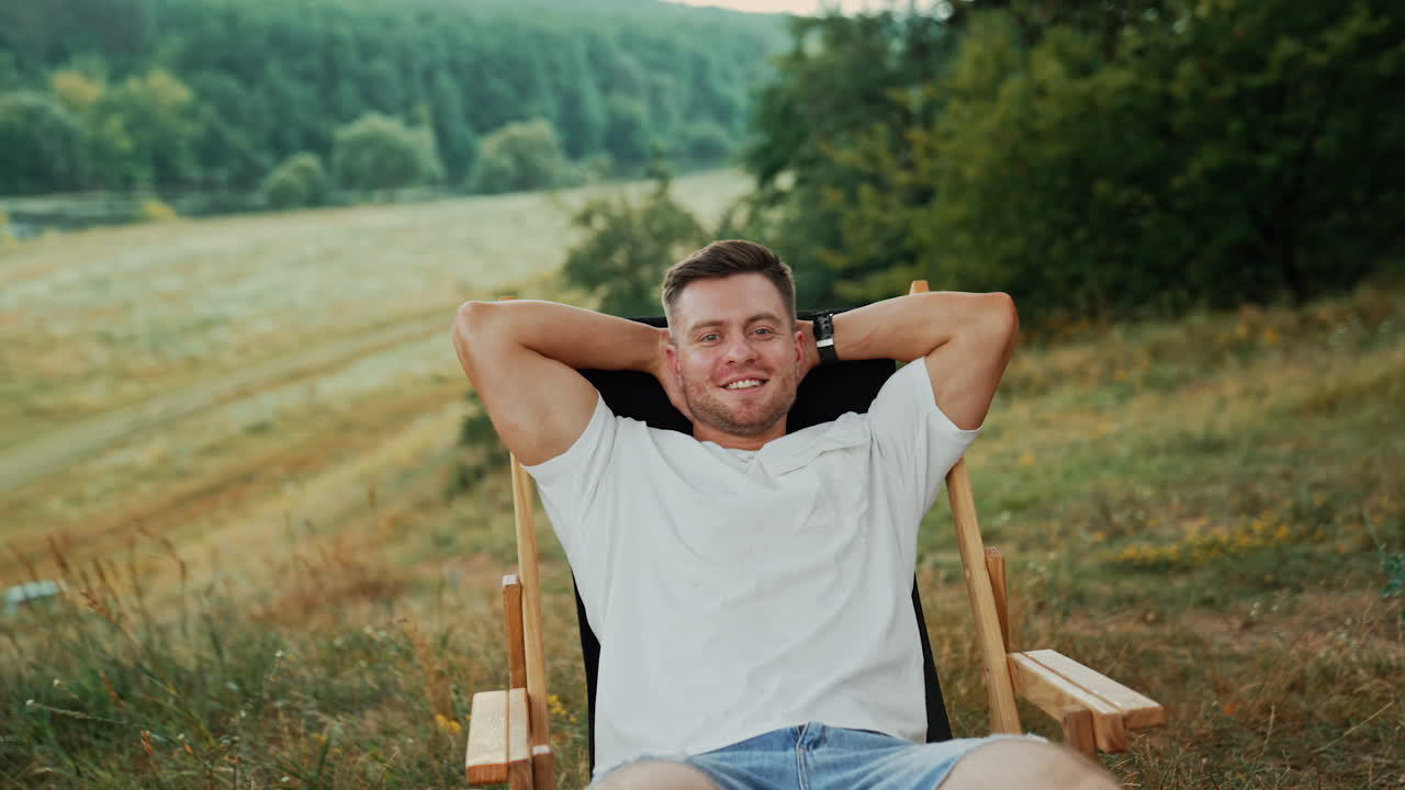 Dreamy Caucasian man sits in a chair his hands behind head. Relaxed man enjoys beautiful nature around him.
