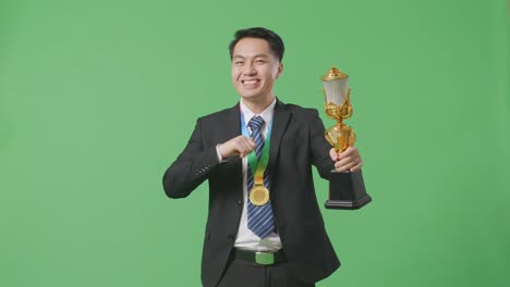 asian business man in a suit and tie with a gold medal showing gold trophy to camera, smiling, and pointing himself being proud winning as the first winner on green screen background in the studio