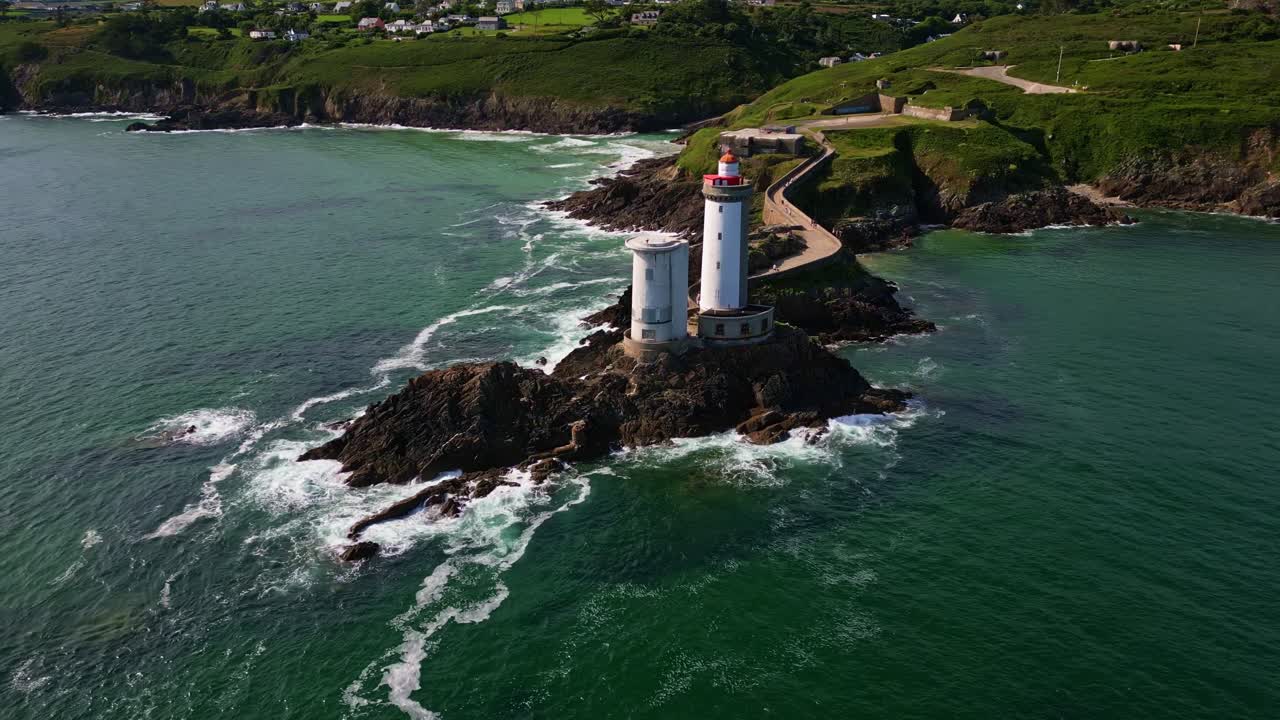 Drone performs a smooth orbit around the Petit Minou lighthouse, showing crashing waves, cliffs, vegetation, and houses under a bright blue sky - Plouzané, Brittany in France