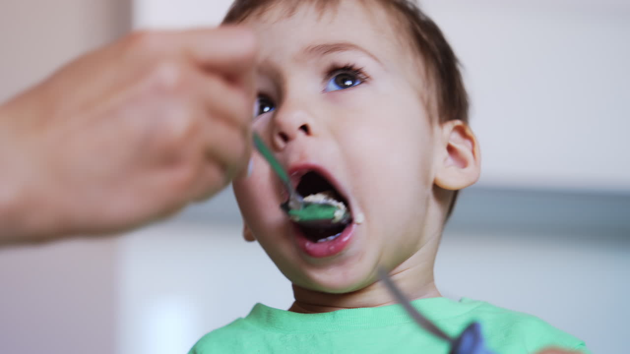 Nice baby boy eating porridge. Kid holds a fork in hand but mom feeds him from spoon. Close up. Blurred backdrop.