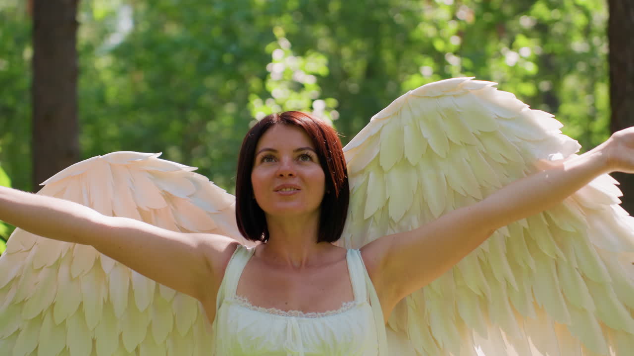 Actress dressed in angelic costume spreads wings and embraces warmth of nature in lush forest as sunlight glows softly around her, creating ethereal atmosphere of peace