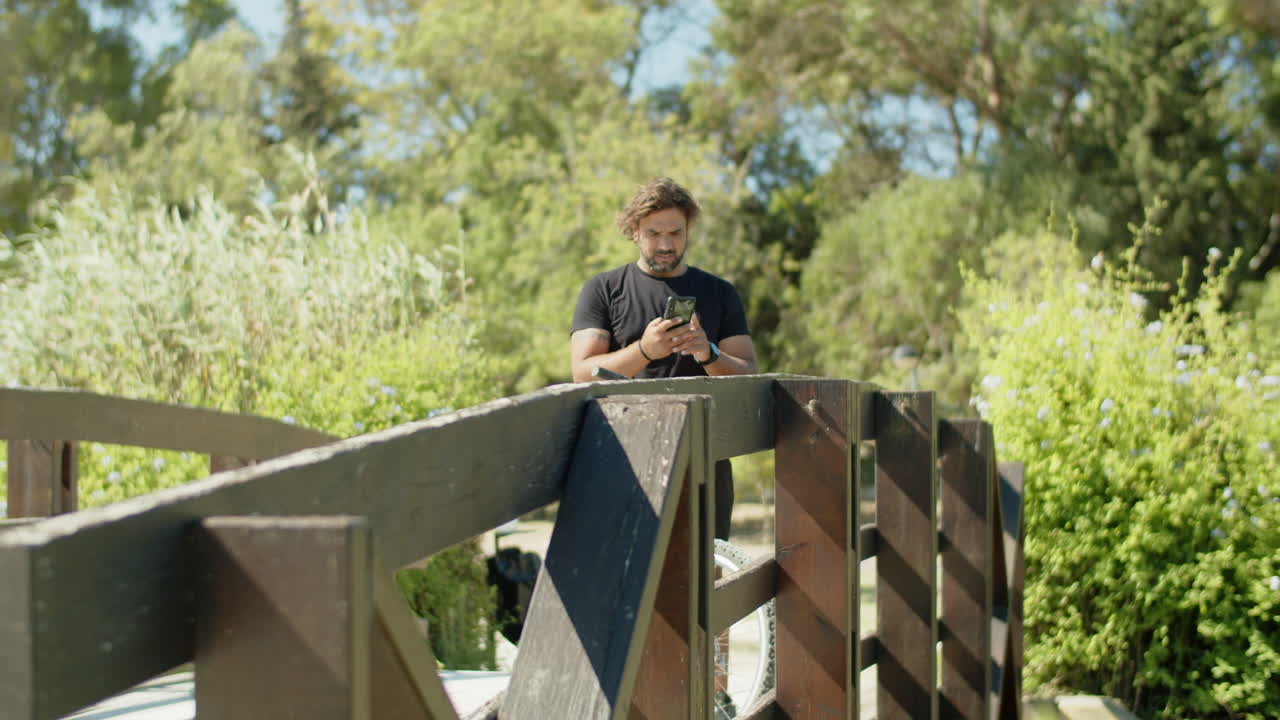 Campo lungo di un uomo in piedi su un ponte con il telefono mentre va in bicicletta.