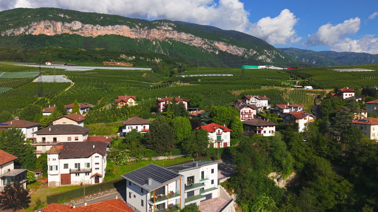 Drone footage of a hillside village surrounded by lush vineyards and rolling hills under a bright summer sky in the Dolomites, Italy