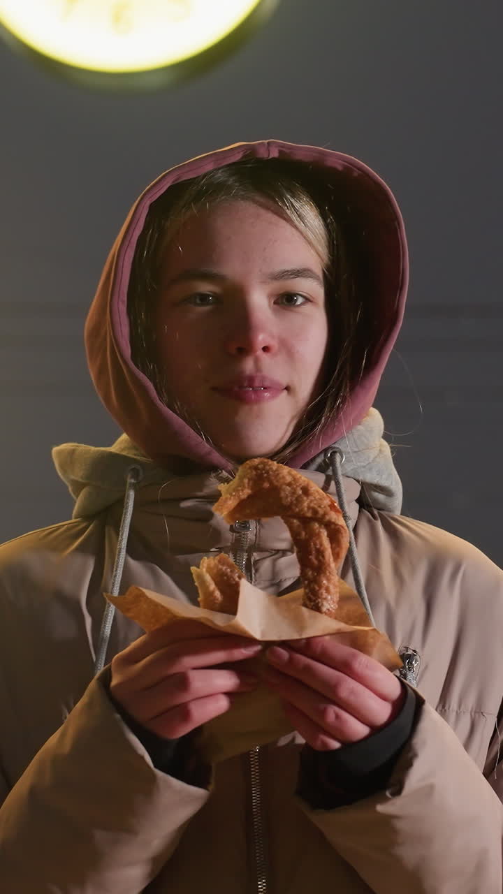 mujer joven con chaqueta de invierno con capucha de pie al aire libre por la noche, con pasteles crujientes en las manos. reloj iluminado en el fondo agrega un brillo cálido al entorno urbano fresco