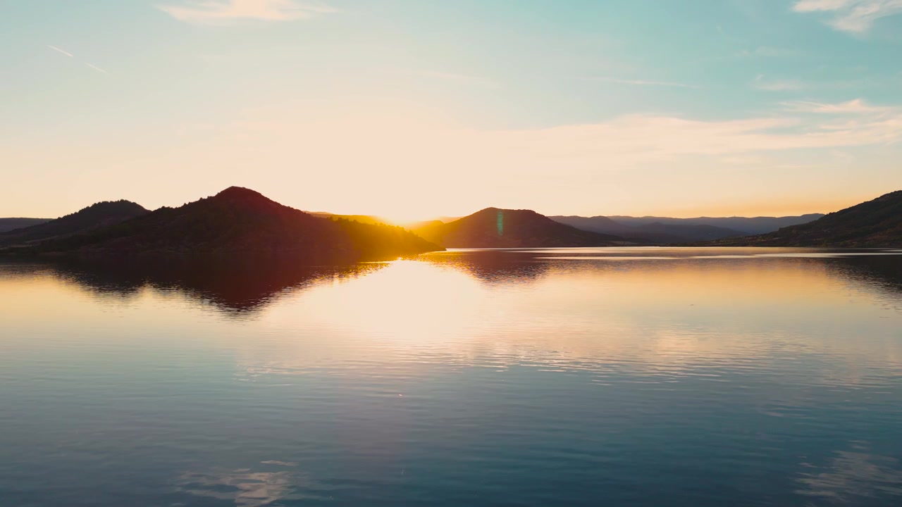 Krásný široký letecký záběr dronem letícím nízko nad dokonale klidnou, skleněnou hladinou jezera Salagou (Lac du Salagou) v Héraultu, zachycující symetrický odraz zvlněných kopců při zlatém západu slunce.