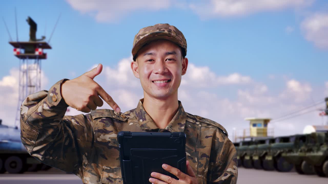아시아인 군인이 미소 짓고 군 캠프에 서 있는 동안 손에 든 태블릿을 가리키고 있습니다.