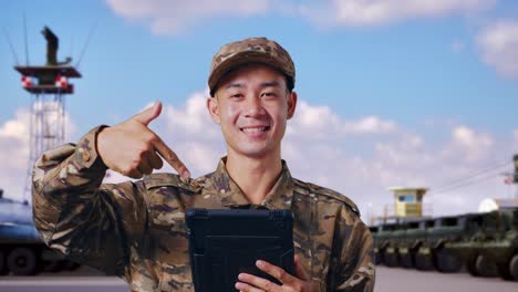 un soldado asiático sonriendo y señalando una tableta en su mano mientras estaba de pie en un campamento militar