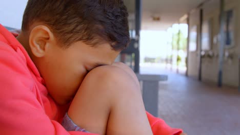 vista lateral de un escolar asiático con la cabeza hacia abajo sentado solo en un banco en el corredor de la escuela 4k