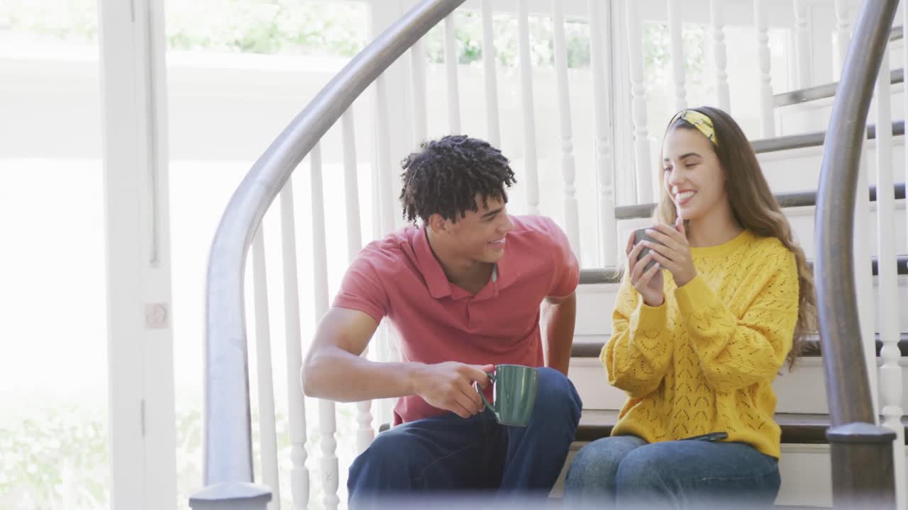feliz pareja biracial pasando el tiempo en casa bebiendo café y sentados en las escaleras