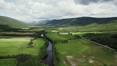 Vista-Aérea-Del-Río-Que-Fluye-A-Través-De-Los-Campos-Verdes-Con-Montañas-Durante-El-Día