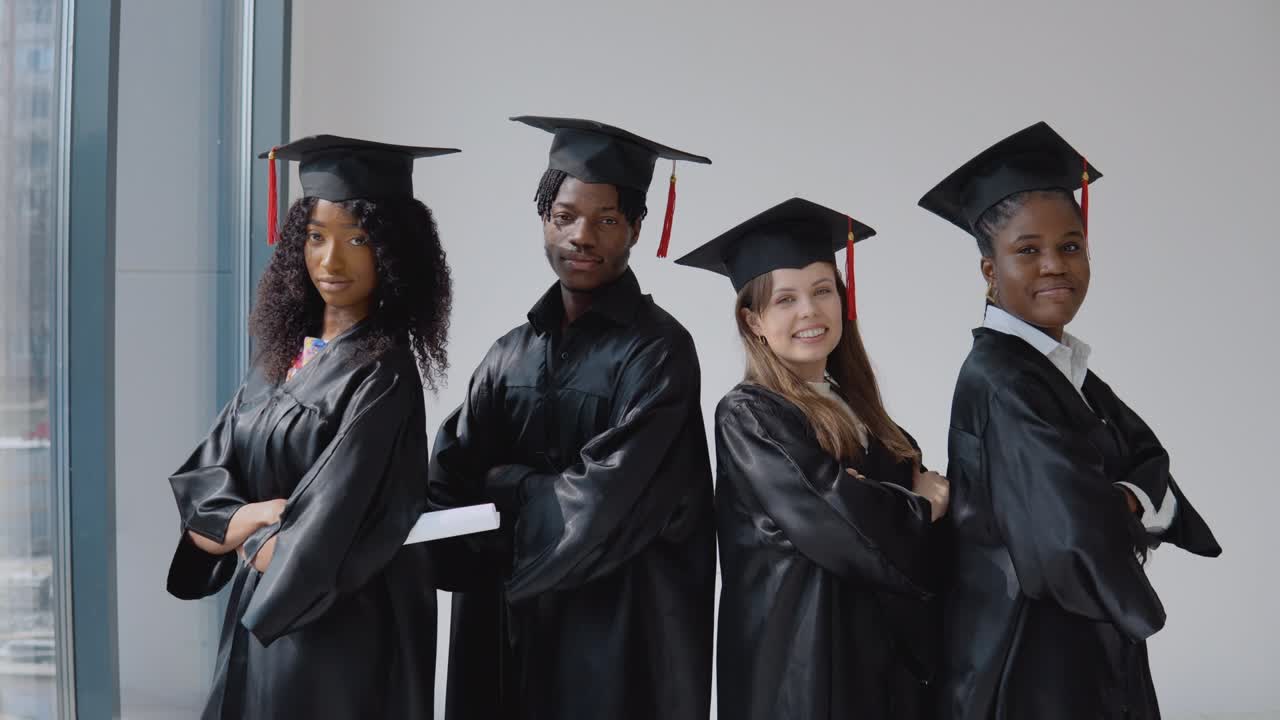 dos mujeres jóvenes y un joven afroamericano y una joven caucásica graduada de la universidad con diplomas y libros en las manos están hombro a hombro junto a la ventana.