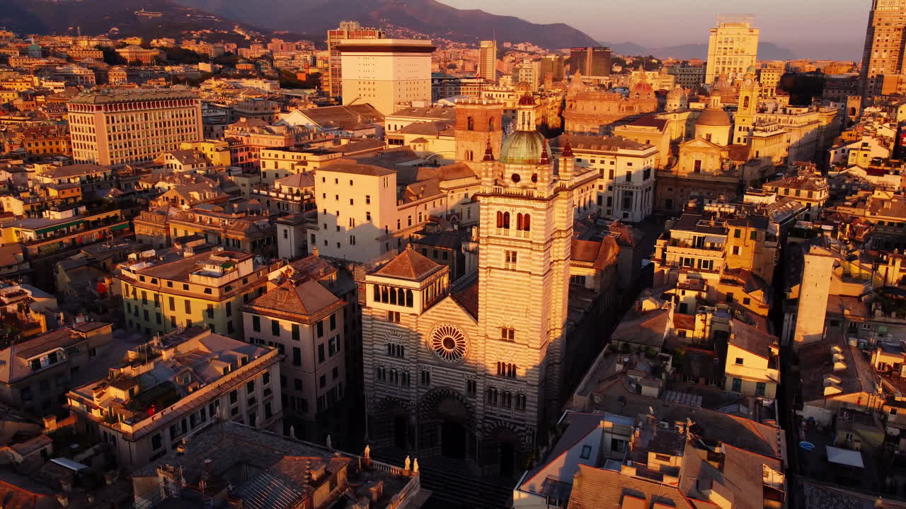 Drone orbiting around Genoa Cathedral in the historical center at sunset, with surrounding old buildings and the cityscape bathed in golden light