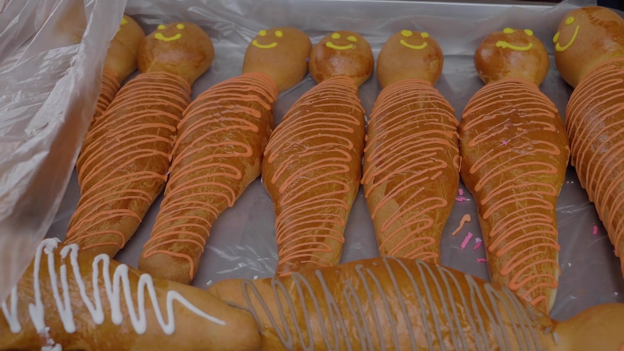 Un primo piano mostra le "Wawas de Pan", un pane tradizionale preparato per il Día de los Muertos, con facce disegnate sopra e glassa a zigzag. Appena sfornato, questo dolce evidenzia il significato culturale