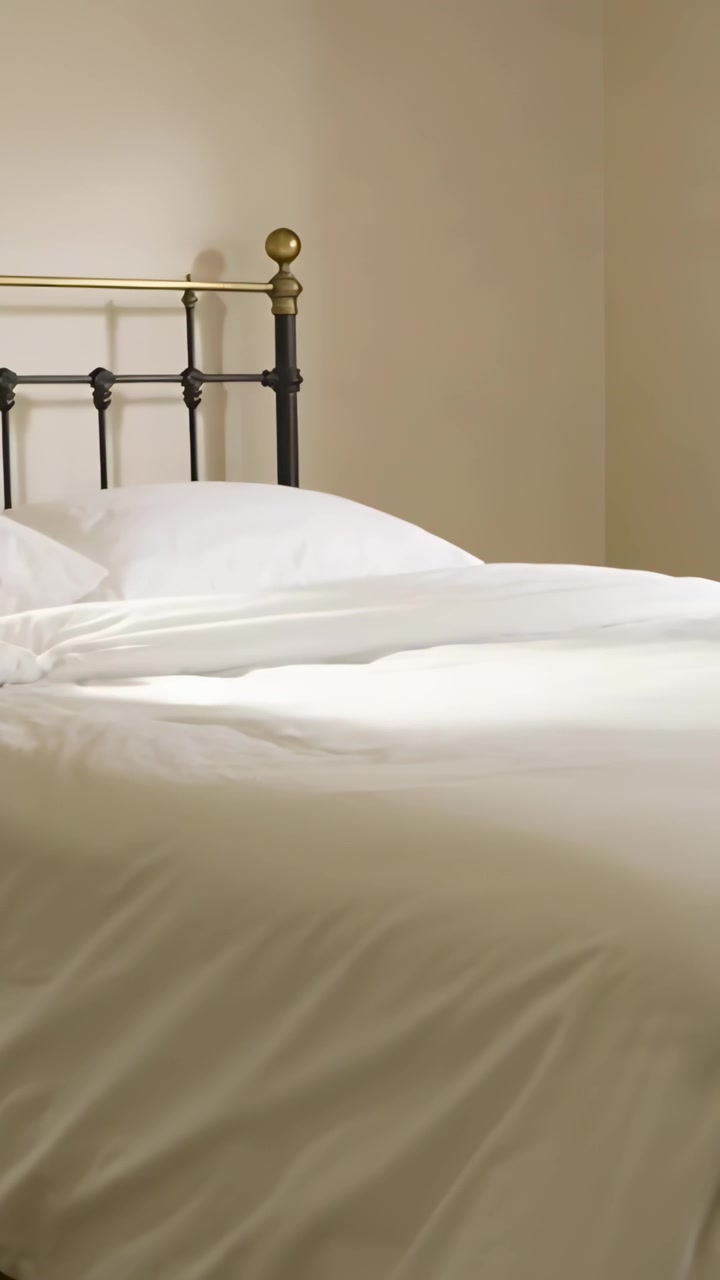 Vertical video: Reaching forearm adjusting bedding on bed in bedroom, small brown dog entering