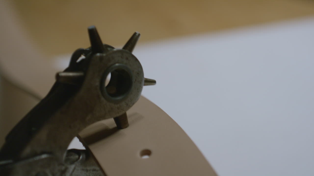 A detailed close-up of a rotary hole punch tool creating precise holes along a curved leather strap on a white surface.