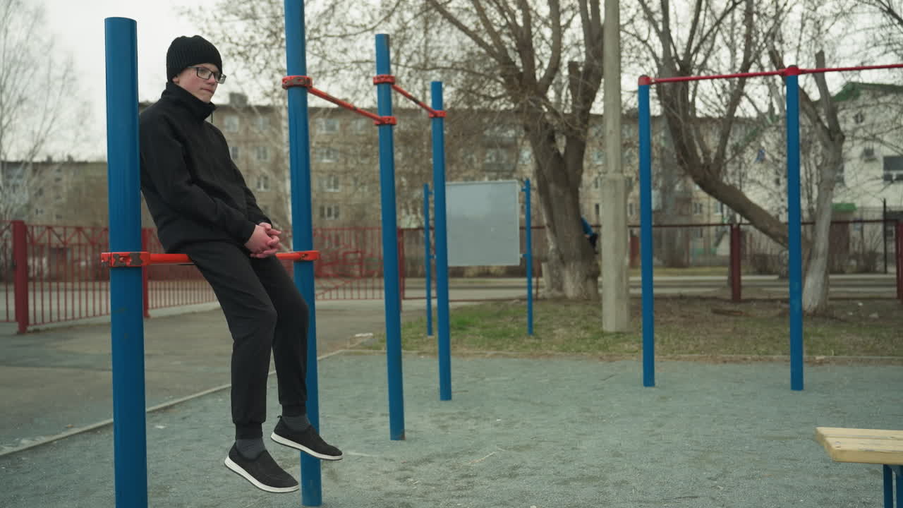 A young boy dressed in black, wearing glasses, sits thoughtfully on a red iron bar, slowly swinging his legs, in the background, a smaller child is seen dropping from a tree and walking away