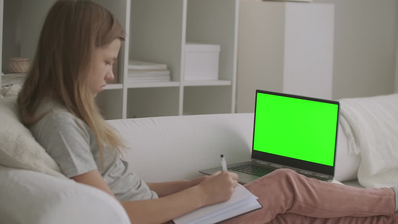 school girl is viewing online lesson on laptop with green screen for chroma key technology,writing at exercise book, learning from home