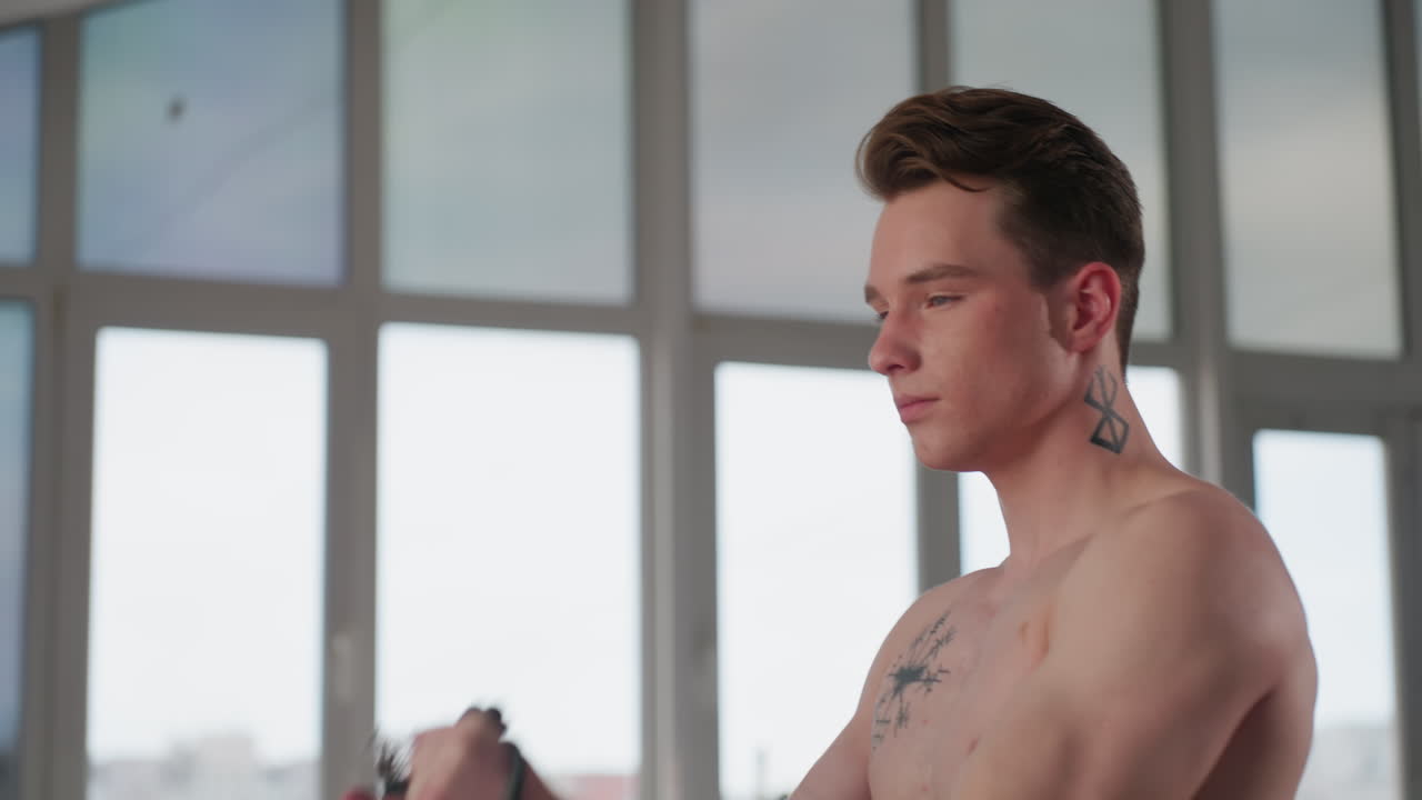shirtless student grips resistance rope with intensity during muscle build up session inside modern space, sunlight pouring through large windows in background