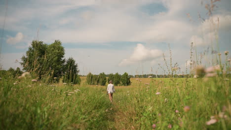 woman in white short walks through tall grassy field holding something in her hand, scene is set under bright sky with scattered clouds, surrounded by green plants and flowers