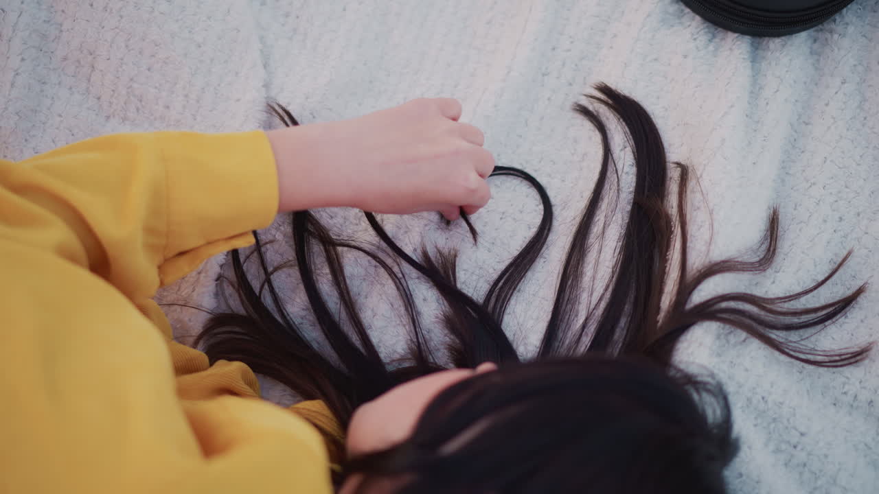 아시아 여성이 니트 담요 위에 누워 있고, 긴 흑발은 손가락으로 부드럽게 가닥을 정리하여 하트 모양을 이루고 있으며, 노란색 스웨터 소매가 보이고, 부드러운 파스텔 톤, 아늑한 로파이 분위기, 친밀하고 조용한 순간