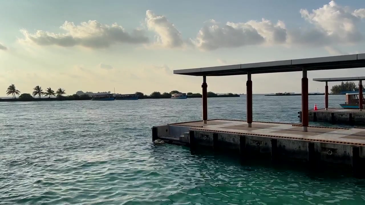 Concrete pier with roof structure extending over ocean water with distant palm trees Maldives