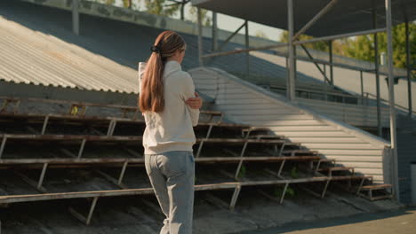 lady in hood and jeans with long hair tied back walking near empty stadium seating on sunny day, hands folded, looking reflective and deep in thought, surrounded by empty bleachers and green trees