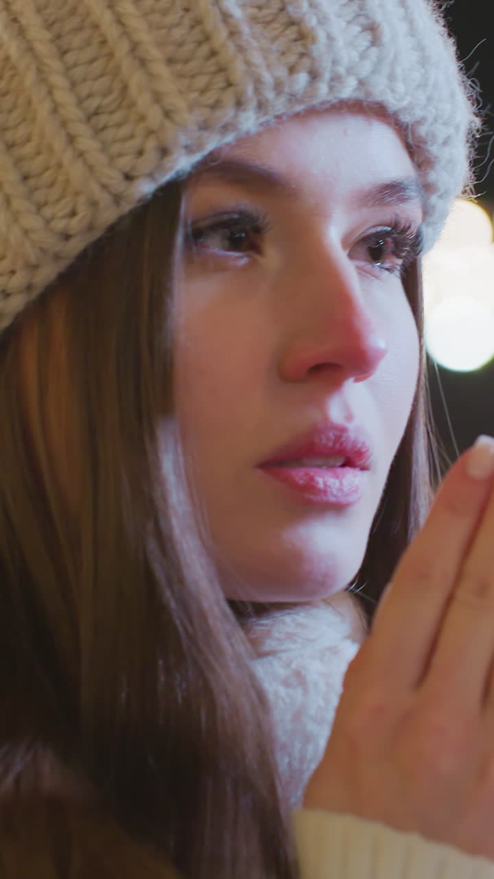 Close-up of woman bringing elegant hands to mouth, exhaling warm breath in cold winter night, visible breath vapor, blurred background features passerby and festive golden lights