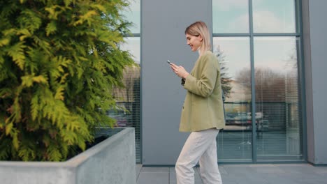 side view. smiling young blonde woman texting messages on smartphon while walking down the city street