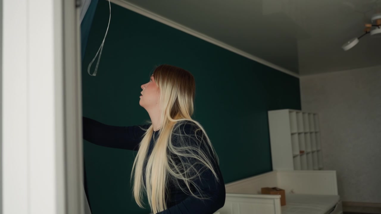 Donna caucasica sistema le tende nel nuovo appartamento, tira indietro la tenda velata e il drappeggio, ispeziona il lampadario e la posizione del comò, stanza vuota con parete d'accento color verde acqua, organizza la disposizione, testa luce e posizionamento