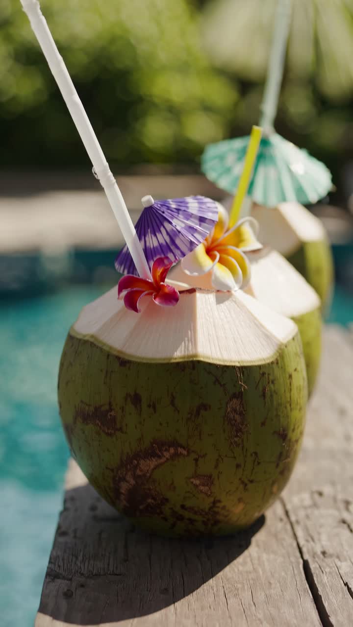 Close-up, eye-level shot of coconuts with colorful umbrellas and straws by a pool