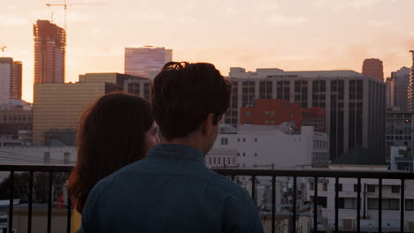 rear view of couple on rooftop terrace looking out over city skyline at sunset