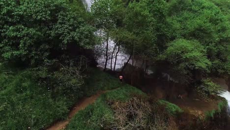 Caucasian-man-stands-at-edge-of-powerful-cascading-waterfall,-aerial-rising-pull-back