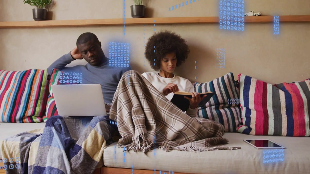 Couple sitting sofa, woman tapping tablet causing HUD graphics rising over laptop fading for tech