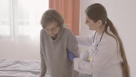 female doctor helps elder woman to get out of bed