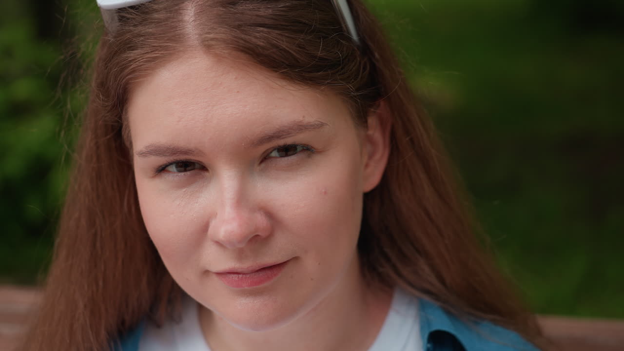 Close up of elegant woman with long brown hair and white sunglasses resting on head giving charming confident look while sitting outdoors on park bench surrounded by lush greenery