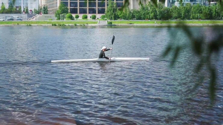 Solo paddler powering forward stroke in narrow white racing kayak on city river, wide water view, grassy embankment and promenade under bright sunlit sky morning