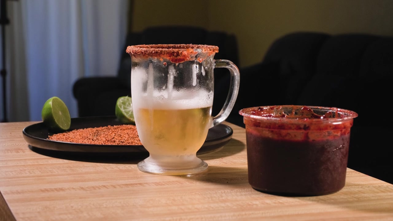 Un uomo ripone un bicchiere di plastica brinato riempito con salsa chamoy scura su un tavolo da cucina accanto a ingredienti freschi.