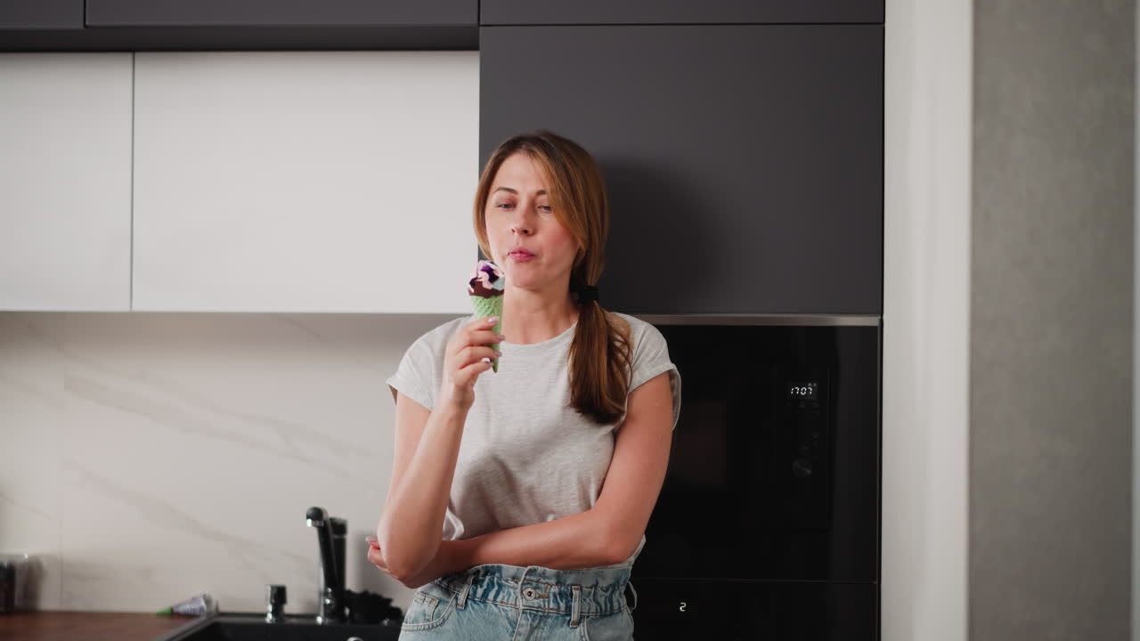 Woman standing relaxed beside fridge in modern kitchen, licking ice cream cone with eyes closed in moment of enjoyment, arms folded, casual outfit, peaceful indoor scene with soft natural light