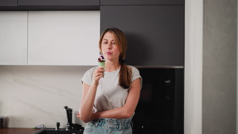 woman standing relaxed beside fridge in modern kitchen, licking ice cream cone with eyes closed in moment of enjoyment, arms folded, casual outfit, peaceful indoor scene with soft natural light