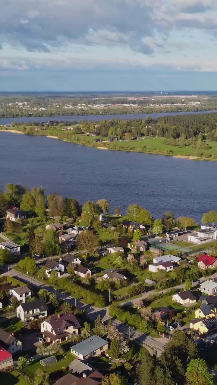 Slow drone flyover shows Katlakalns residential area along Daugava River right bank with tree-lined streets, garden houses, opposite Dole Island forest, open fields, Gulf of Riga visible in distance